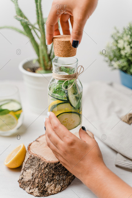 Crop woman opening glass jar of tasty lemonade with slices of fresh lemon and cucumber with green leaves of mint on wooden tray while working at white table