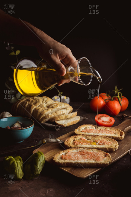 Unrecognizable cook pouring oil on pieces of fresh bread with sauce while preparing toasts on black background