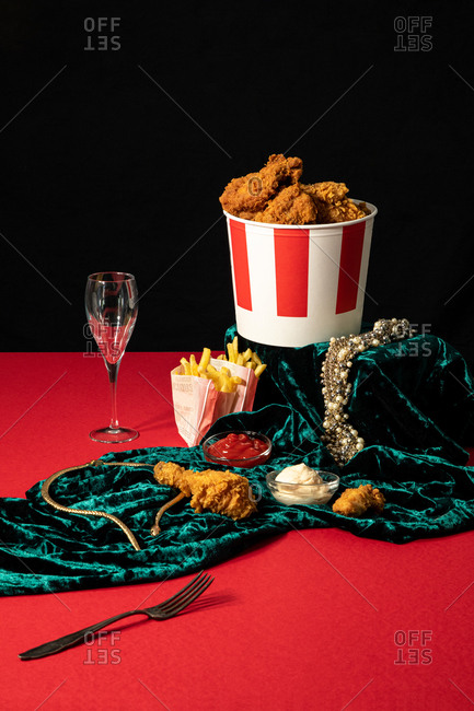Deep fried chicken and portions of french fries in composition with sauces and golden jewelry on shiny green velvet on red table on background of black wall