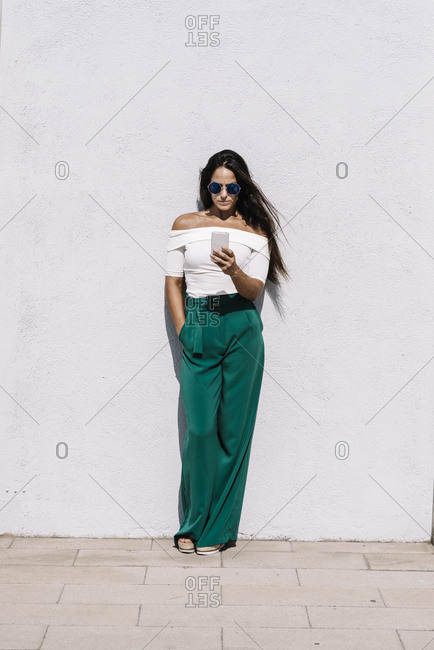 Beautiful long haired woman with sunglasses leaning on a white wall while using a mobile phone in a sunny day