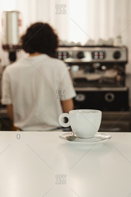 White cup of coffee with saucer and spoon placed on coffee shop counter with blurred female barista using coffee machine in background