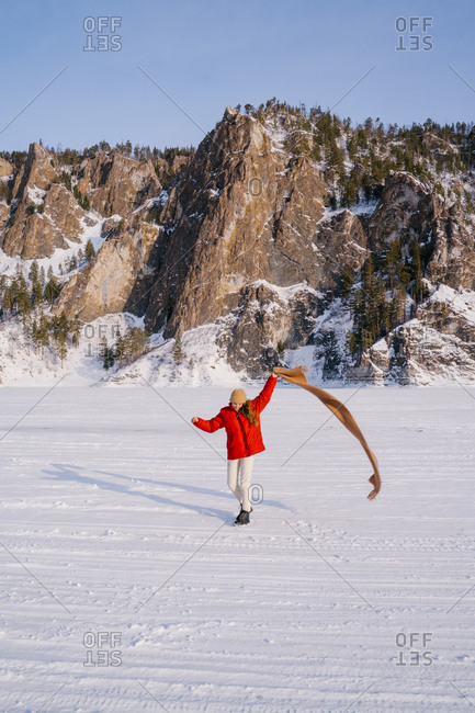 Happy female in red jacket running with scarf in raised hand on snowy valley with majestic rocks on background and enjoying sunny winter day in Siberia Russia