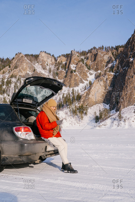 Side view of female in warm active wear sitting on open car trunk and using phone in snowy valley with beautiful rocks during winter road trip in Siberia Russia