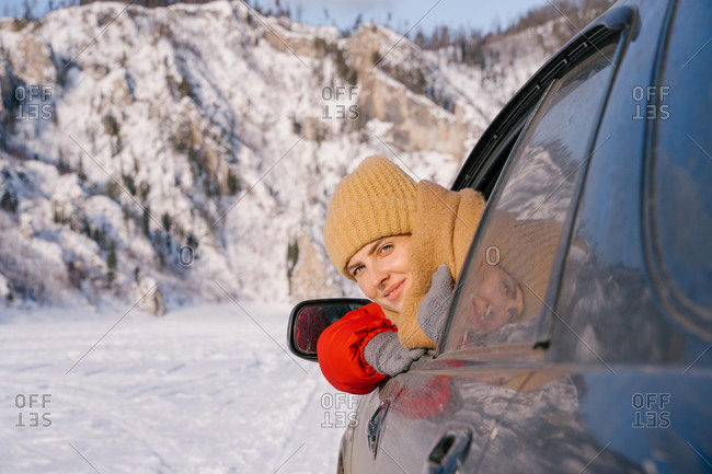 Happy smiling young female traveler in warm clothes sitting in car and leaning out window during road trip to snowy mountains in Siberia Russia