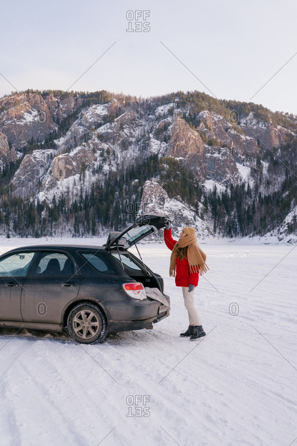 Side view of active woman in warm clothes opening car trunk while standing in snowy valley next to majestic rocks in Siberia Russia