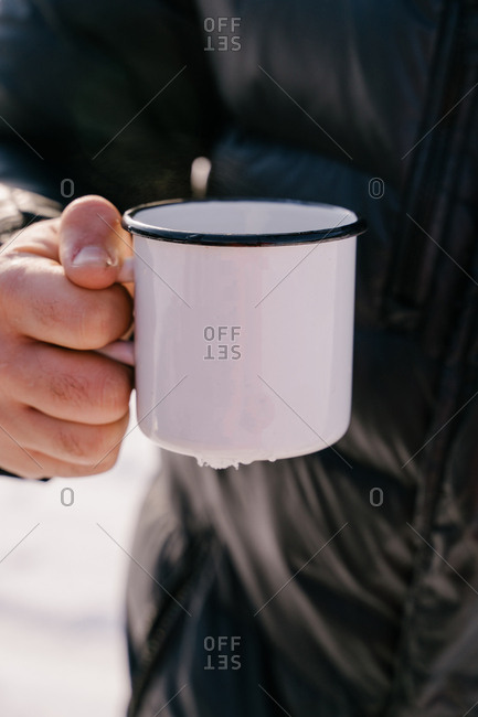 Crop male traveler in warm black jacket holding white enamel mug with hot tea while standing outdoors on winter day in Siberia