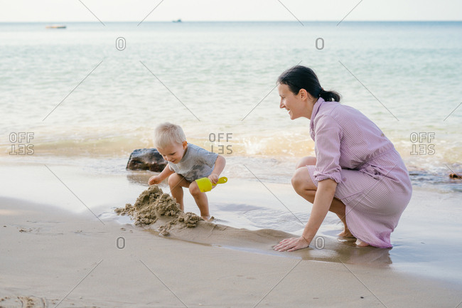 Toddler and mother playing with sand on beach against blurred seascape in sunny day