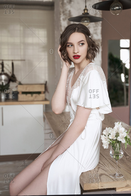 Beautiful tender woman in white silk dressing gown touching hair and pondering while sitting on table beside flowers in wineglass and looking away in apartment
