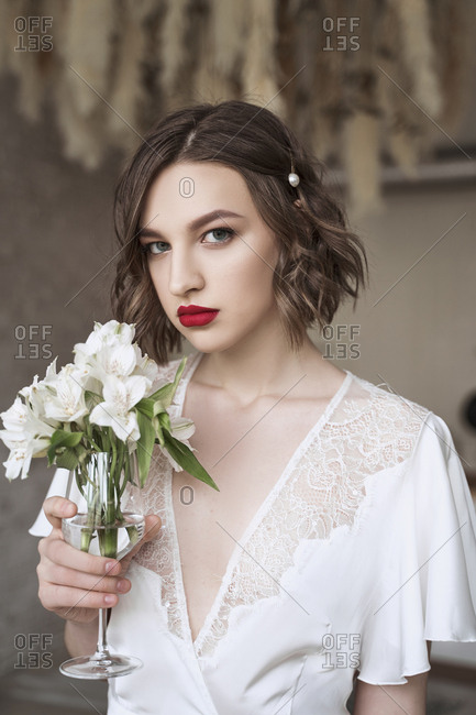 Charming modest lady in white dress with lace looking away and dreaming while holding glass with small white flowers and enjoying scent