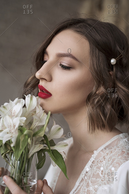 Charming modest lady in white dress with lace looking away and dreaming while holding glass with small white flowers and enjoying scent