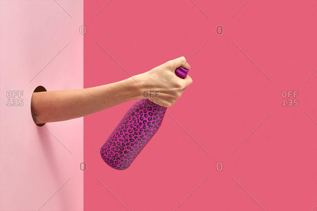 Female's hand holds painted black spots wine bottle closed by the thumb through the hole in the wall on a duotone pink background with soft shadows, copy space.