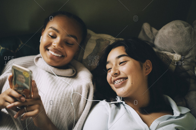 Happy female teenagers listening music on smart phone while lying on bed at home