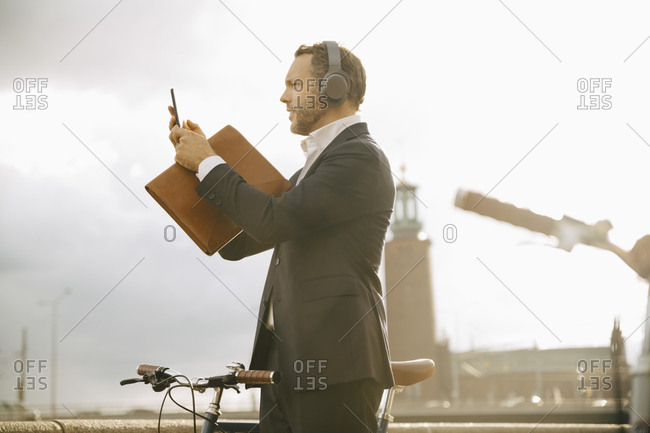 Side view of businessman photographing through mobile phone while standing by bicycle against sky in city