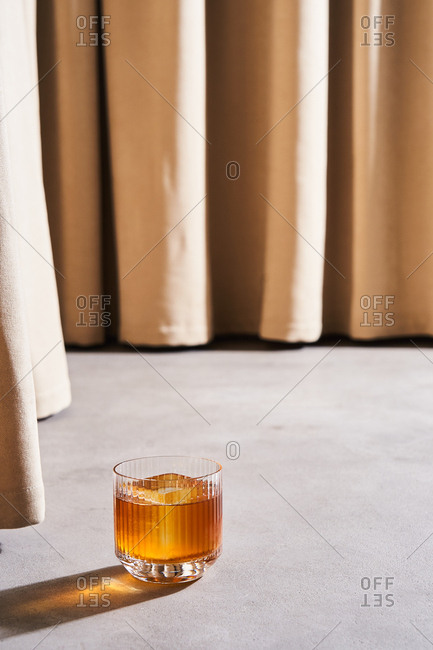 Golden colored cocktail with large ice cubes by beige drapes