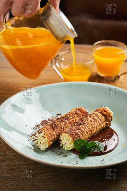 Person pouring orange juice beside dish with delicious cannolis
