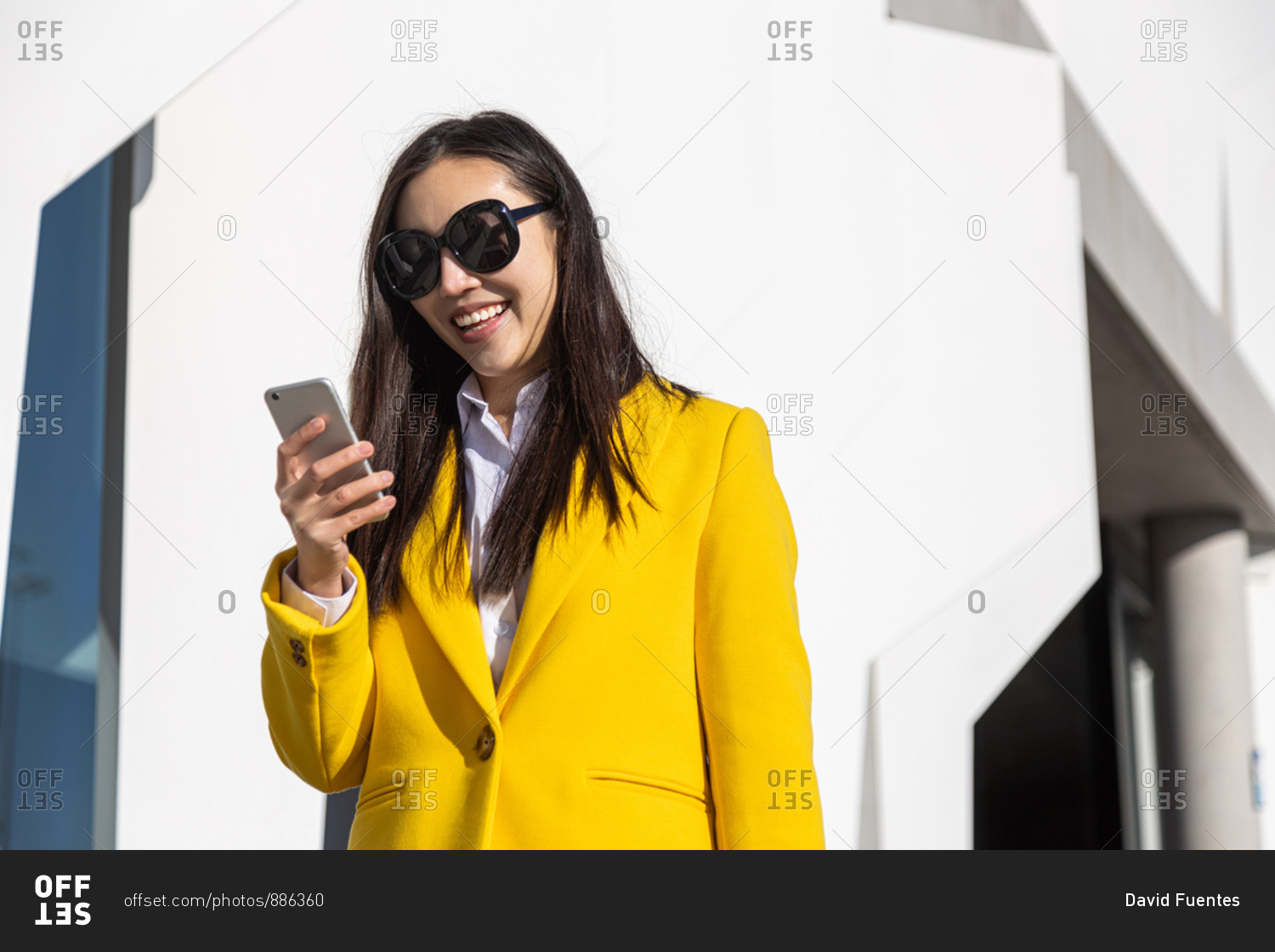 Smiling Asian business woman with yellow coat using cell phone with building in the background
