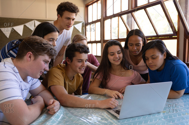 Front view of a multi-ethnic group of teenage school pupils sitting in a classroom looking at a laptop computer together and smiling at break time