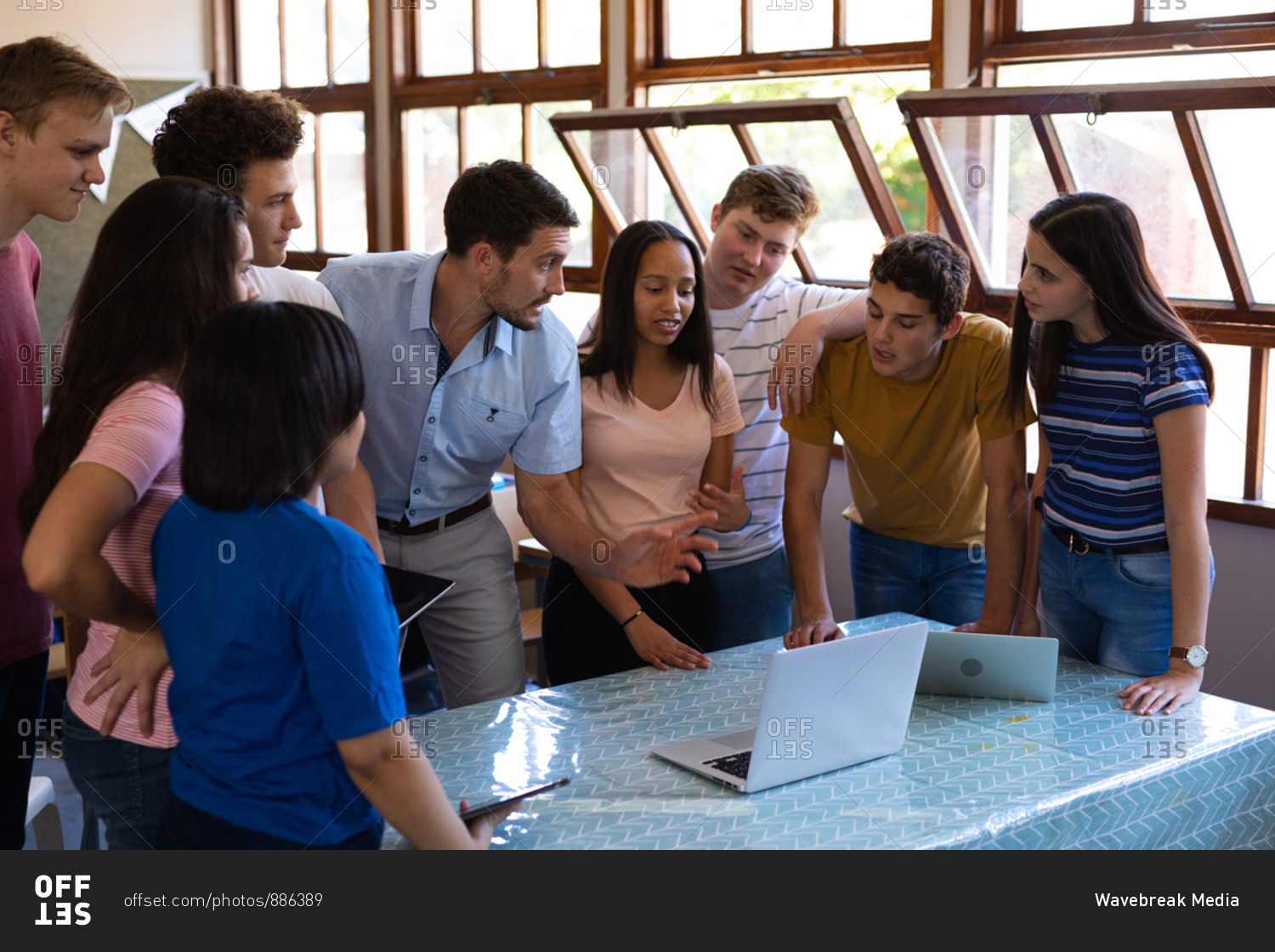 Front view of a multi-ethnic group of teenage school pupils and their Caucasian male teacher standing in a classroom looking at laptop computers together, the pupils listening while the teacher talks