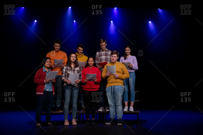 Front view of a multi-ethnic group of teenage male and female choristers holding sheet music and singing standing on the stage of a school theatre during rehearsals for a performance