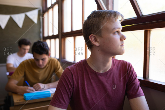 Front view close up of a Caucasian teenage boy sitting at a desk in a school classroom looking out of the window, with classmates sitting at desks working in the background