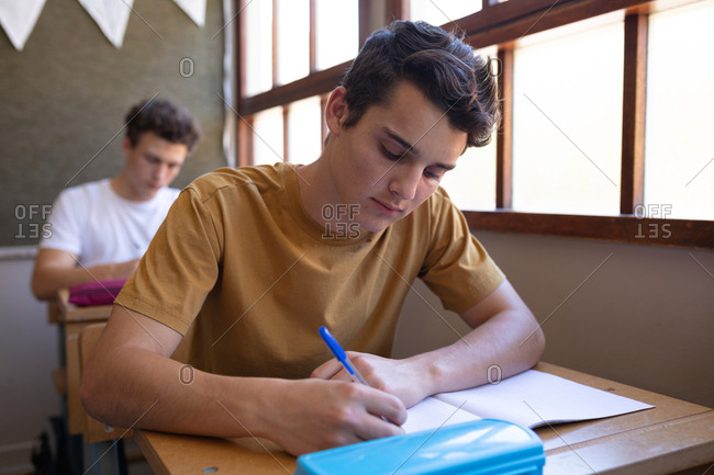 Front view close up of a Caucasian teenage boy sitting at a desk in a school classroom writing in a notebook, with a classmate sitting at a desk in the background