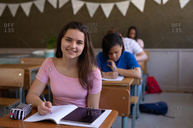 Portrait of a Caucasian teenage girl sitting at a desk in a school classroom writing in a notebook and looking to camera smiling, with classmates sitting at desks working in the background