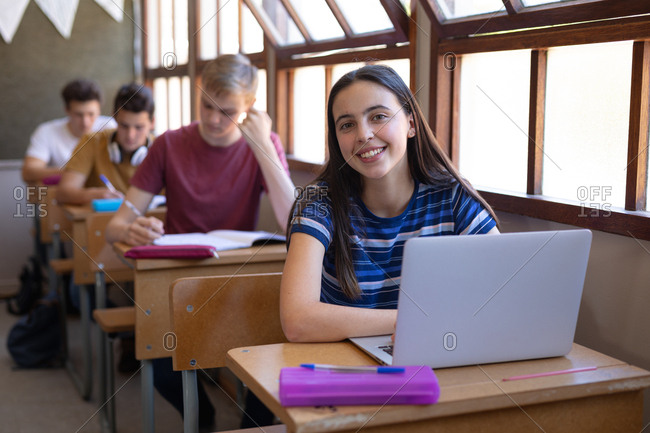 Portrait of a Caucasian teenage girl sitting at a desk in a school classroom using a laptop computer and looking to camera smiling, with classmates sitting at desks working in the background