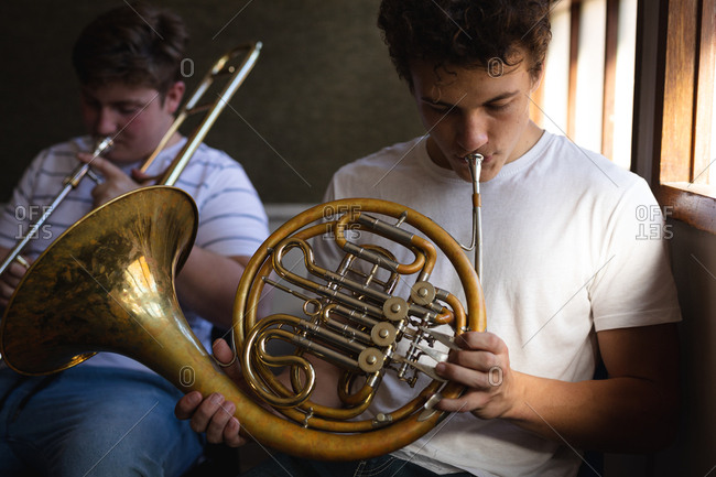 Front view of two Caucasian teenage male musicians rehearsing, one playing the French horn and one playing the trombone. High school education concept.