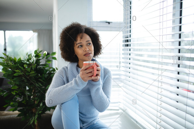 Front view of a mixed race woman relaxing at home, sitting on a window seat looking out of the window, holding a cup of coffee and smiling