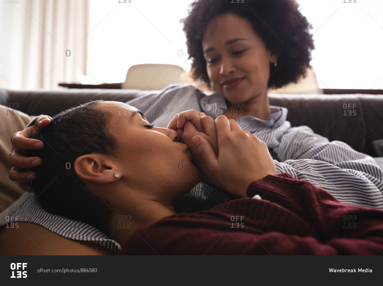 Side view close up of a mixed race female couple relaxing at home in the living room on the couch together in the morning, one woman lying with her head on the lap of her seated partner, holding and kissing her hand