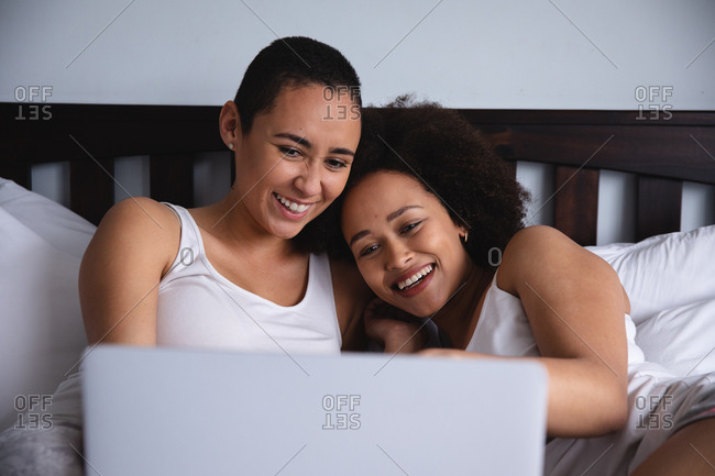 Front view close up of a mixed race female couple relaxing at home in the bedroom, sitting up in bed using a laptop computer together and smiling