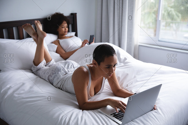 Side view of a mixed race female couple relaxing at home in the bedroom in the morning, one sitting up in bed using a smartphone and the other lying on the bed using a laptop computer