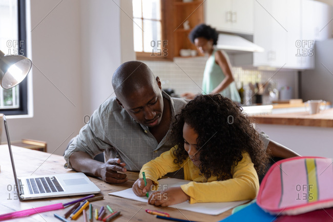 Front view of an African American man at home, sitting at a table with his young daughter watching her drawing in a schoolbook, a laptop computer on the table in front of him, with the mother standing in the kitchen in the background