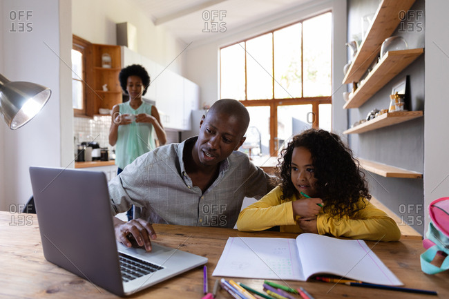 Front view of an African American man at home, sitting at a table with his young daughter looking at a laptop computer together, a schoolbook on the table in front of her, with the mother standing in the kitchen in the background