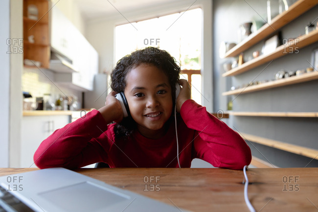 Front view close up of a young African American girl at home, sitting at the dinner table listening with headphones on, plugged into a laptop computer on the table in front of her, looking to camera and smiling