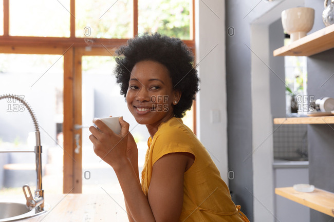 Side view of an African American woman at home, standing in the kitchen holding a cup of coffee with head turned towards the camera, looking away smiling