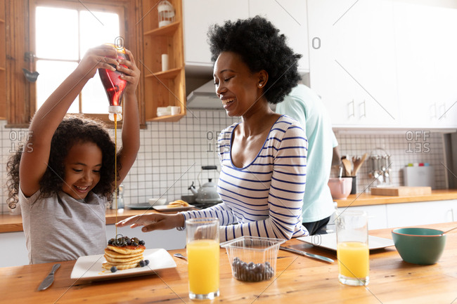 Front view of a young African American girl and her mother at home in the kitchen in the morning, sitting at the kitchen island, the girl pouring sauce on her pancakes and the mother laughing, with the father standing in the background cooking