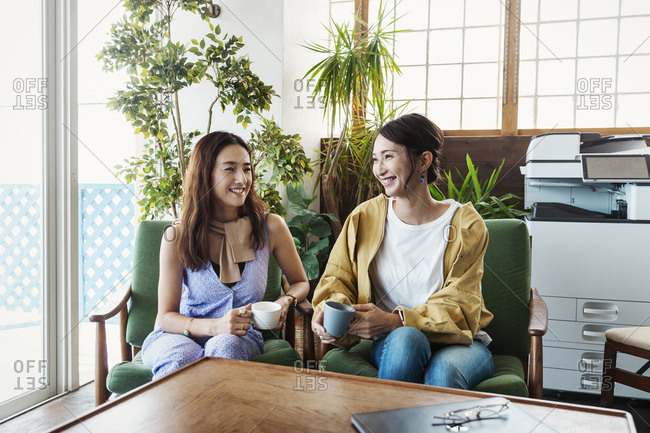 Two female Japanese professionals sitting in a co-working space, smiling at each other.