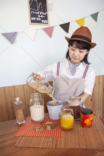 Japanese woman wearing hat standing in a farm shop with a selection of foods and condiments in glass jars.