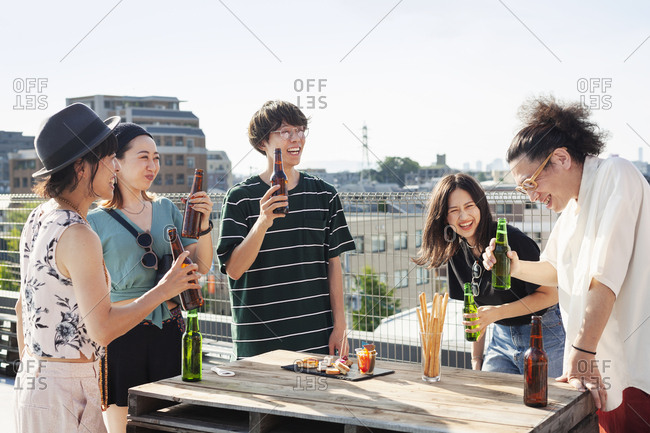 Group of young Japanese men and women standing on a rooftop in an urban setting, drinking beer.