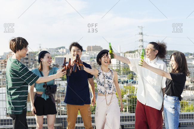 Group of young Japanese men and women standing on a rooftop in an urban setting, drinking beer.