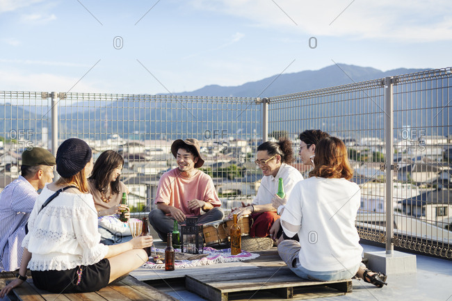 Group of young Japanese men and women sitting on a rooftop in an urban setting, drinking beer.