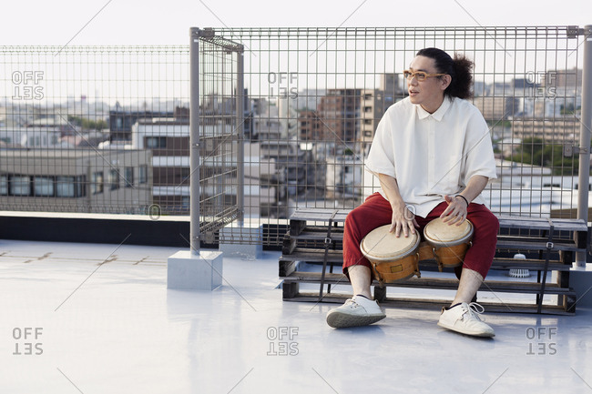 Japanese man sitting on a rooftop in an urban setting, playing drums.