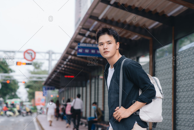 Young handsome man in shirt with hand in pocket and backpack standing near bus stop