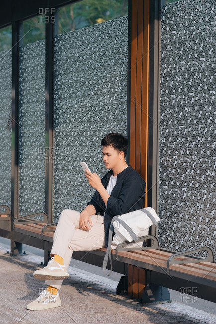 Young handsome asian man using mobile phone while waiting at bus stop