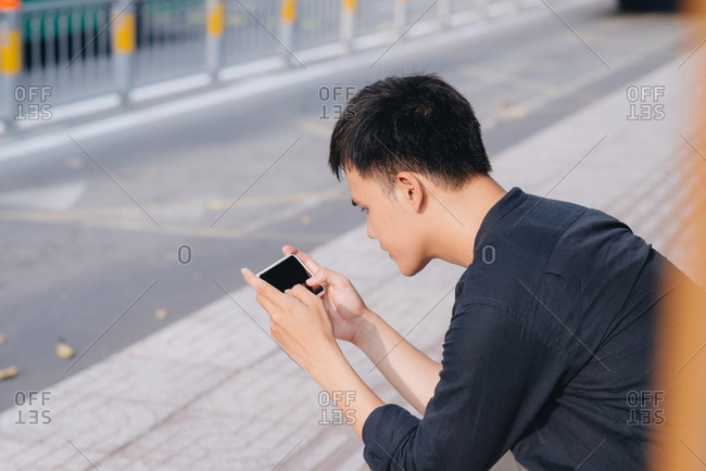 A boy is sitting on the bus stop bench with a cellphone in his hands.