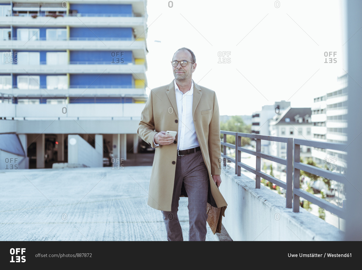 Mature businessman with smartphone and briefcase walking on parking deck
