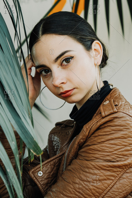 The young beautiful brunette wearing brown leather biker jacket is posing indoor near a green exotic plant