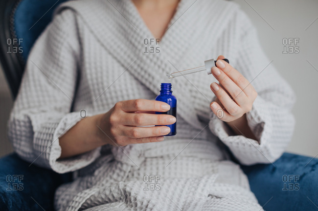 Hands of unrecognizable woman in bathrobe holding bottle of cosmetic serum and a pipette.