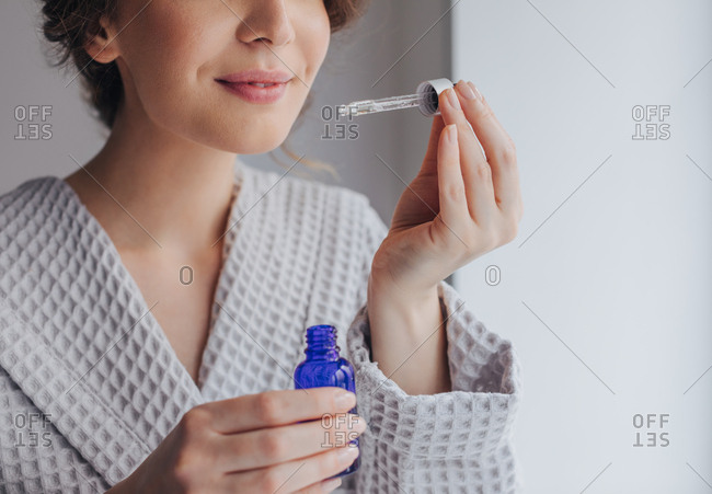 Hands of unrecognizable woman in bathrobe holding bottle of cosmetic serum and a pipette.